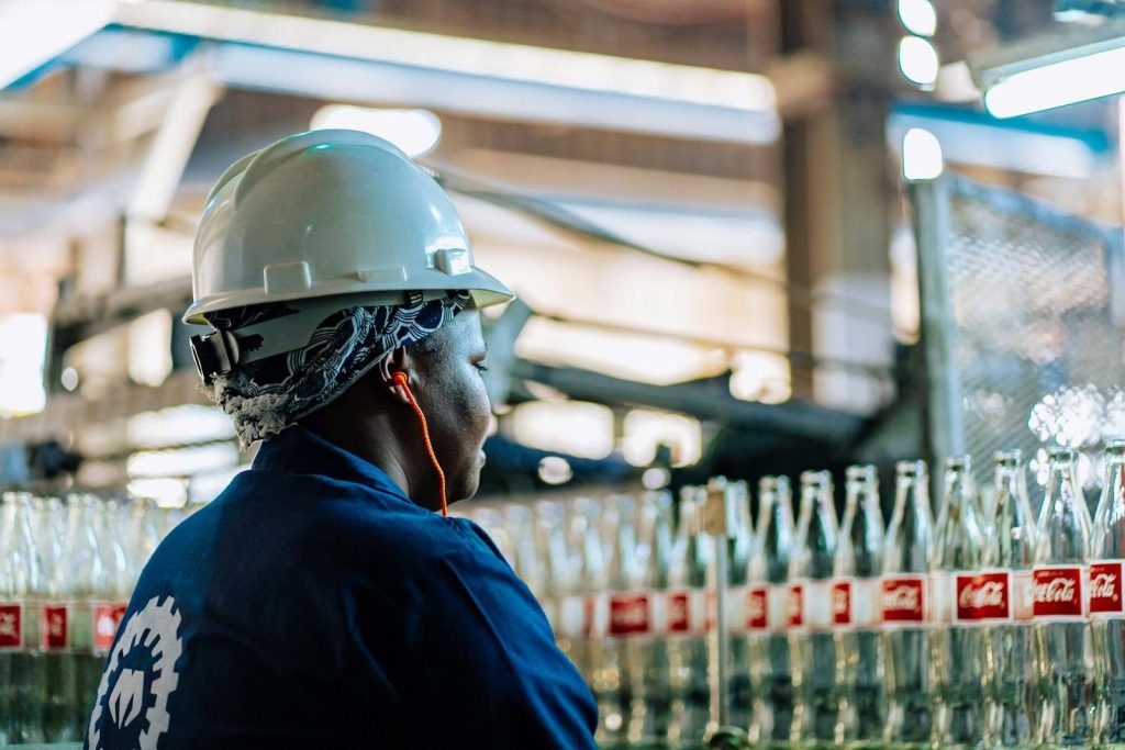 A worker wearing safety gear inspects glass bottles in a factory.