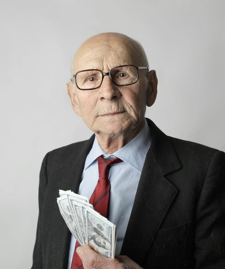 An elderly man with glasses holding dollar bills, representing wealth and financial security.